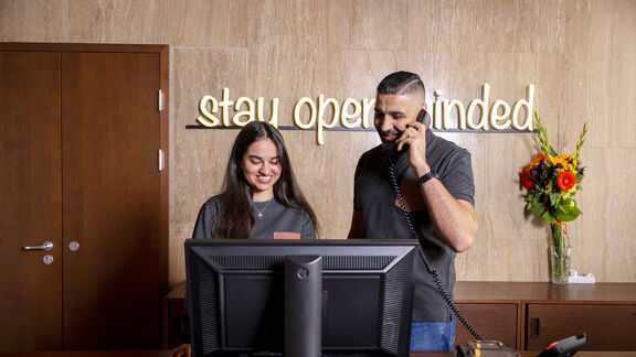 Zwei fröhliche Mitarbeiter:innen stehen an der Rezeption von magdas HOTEL. An der Wand ist ein Schriftzug mit den Worten "stay open-minded" zu sehen.